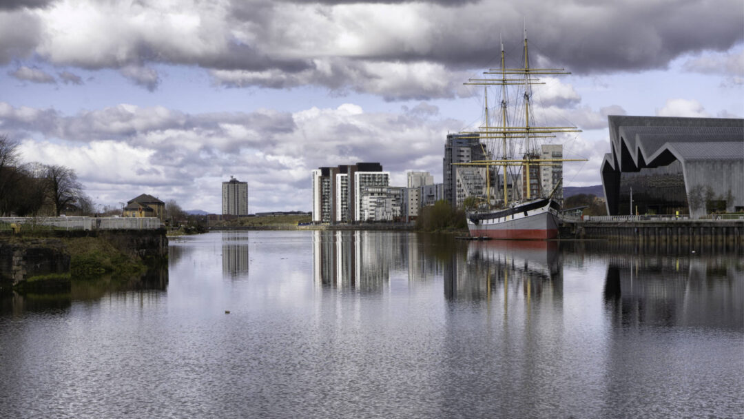A photograph shot from Govan looking up the Clyde towards the Glasgow Transport Museum, in front of which a sailing boat is moored. The reflections in the water create a mirror effect.