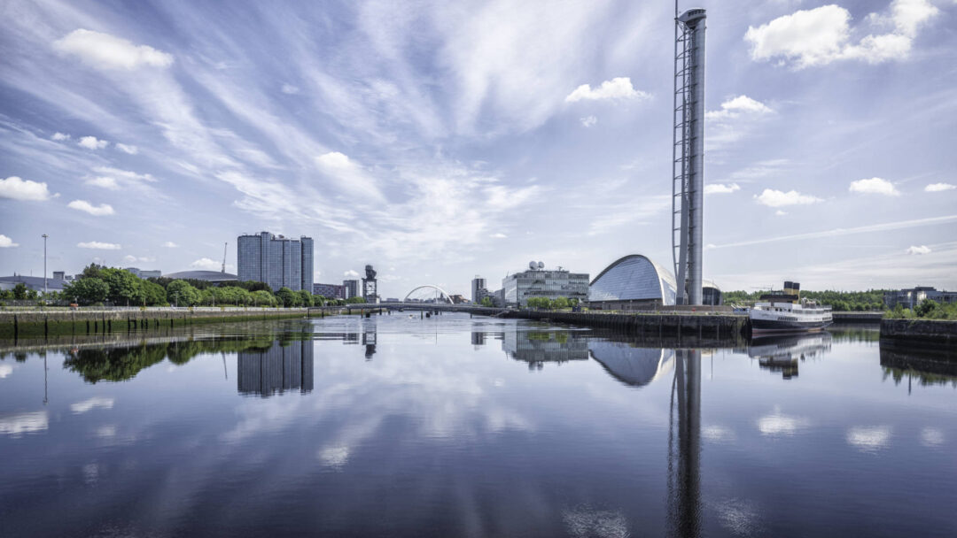 A photograph looking across the Clyde towards the Glasgow Science Centre, BBC Scotland Building and Crowne Plaza Hotel. The sky and water are bright blue, and the reflections on the water create a mirror effect.