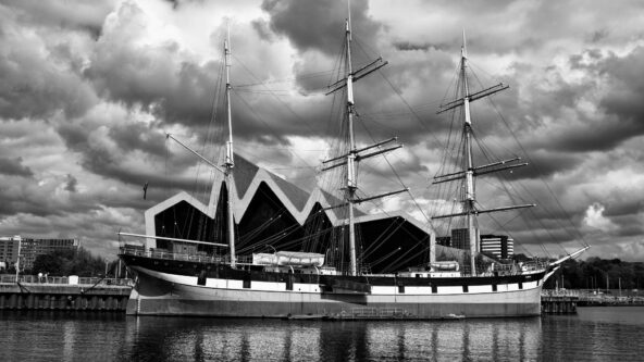 A black and white image, depicting a sailing ship moored in front of the Glasgow Transport Museum on the Clyde, while dark clouds roll above.