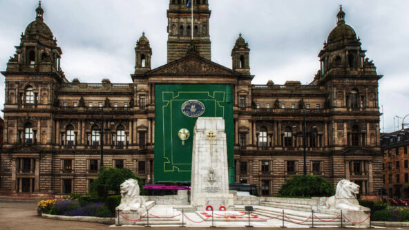 A photograph taken from the East end of George Square. It depicts the Cenotaph monument against the backdrop of the Glasgow City Chambers.